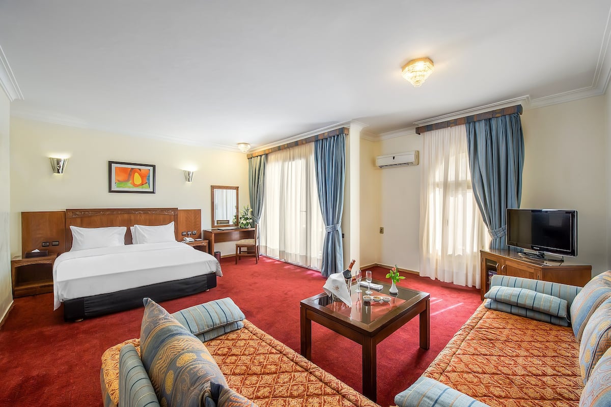A hotel room with a large bed, a sofa set, a television, and a desk with a lamp.