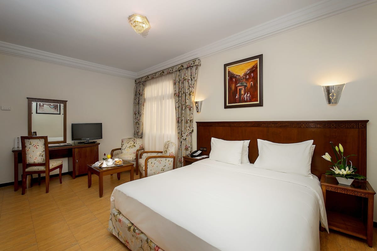 A hotel room with a large bed, a desk with a television, a chair, and a nightstand with a lamp.