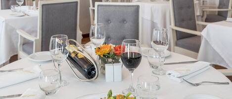 A dining area with white tables, chairs, and a chandelier. There are framed pictures on the walls and a menu stand with a bottle of wine.
