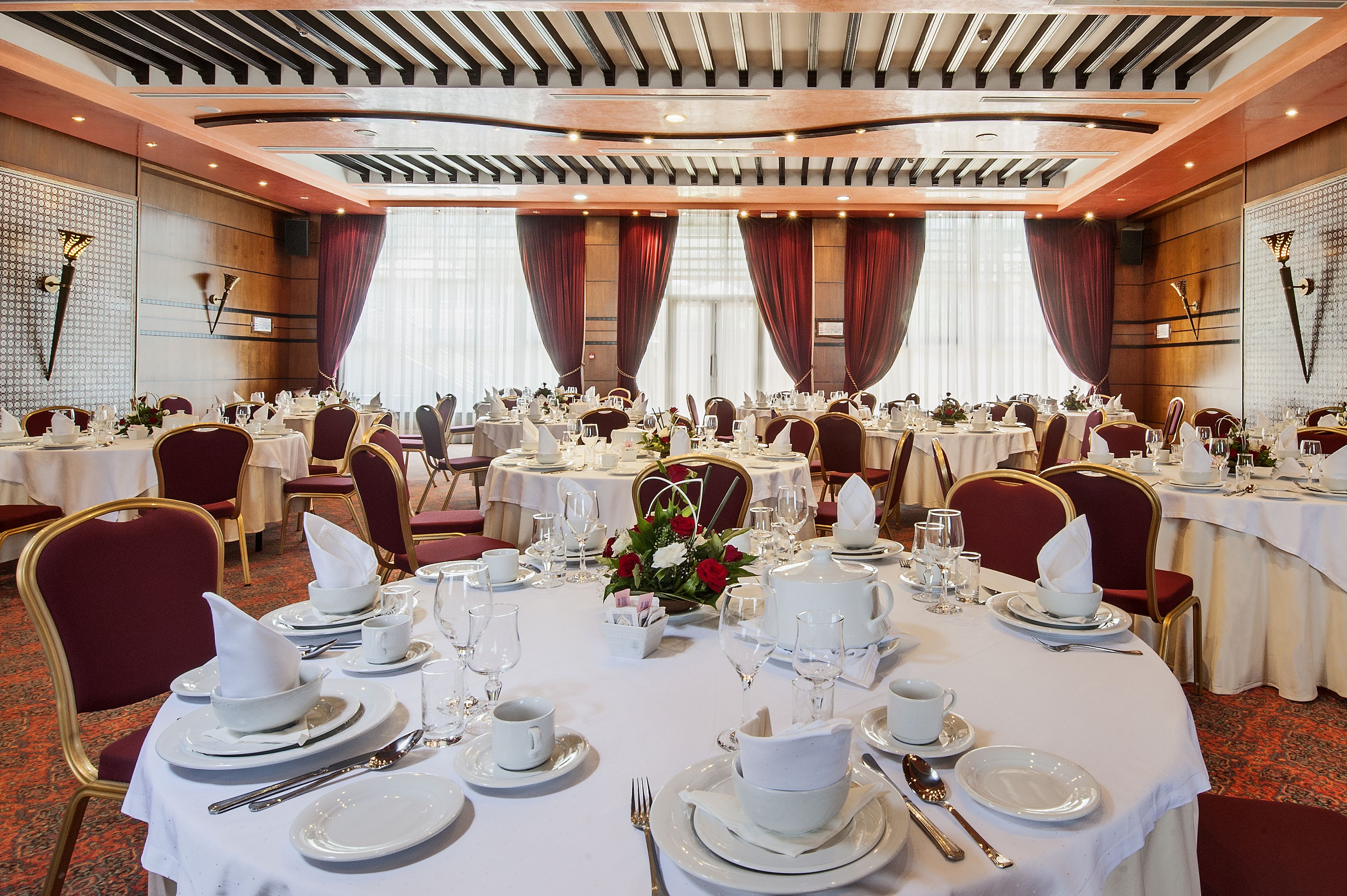 A banquet hall with round tables set for dining, featuring white tablecloths, gold chairs, and red curtains.