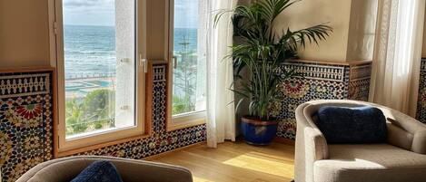 A room with a view of the ocean, two armchairs, a potted plant, and a tiled wall with a geometric pattern.