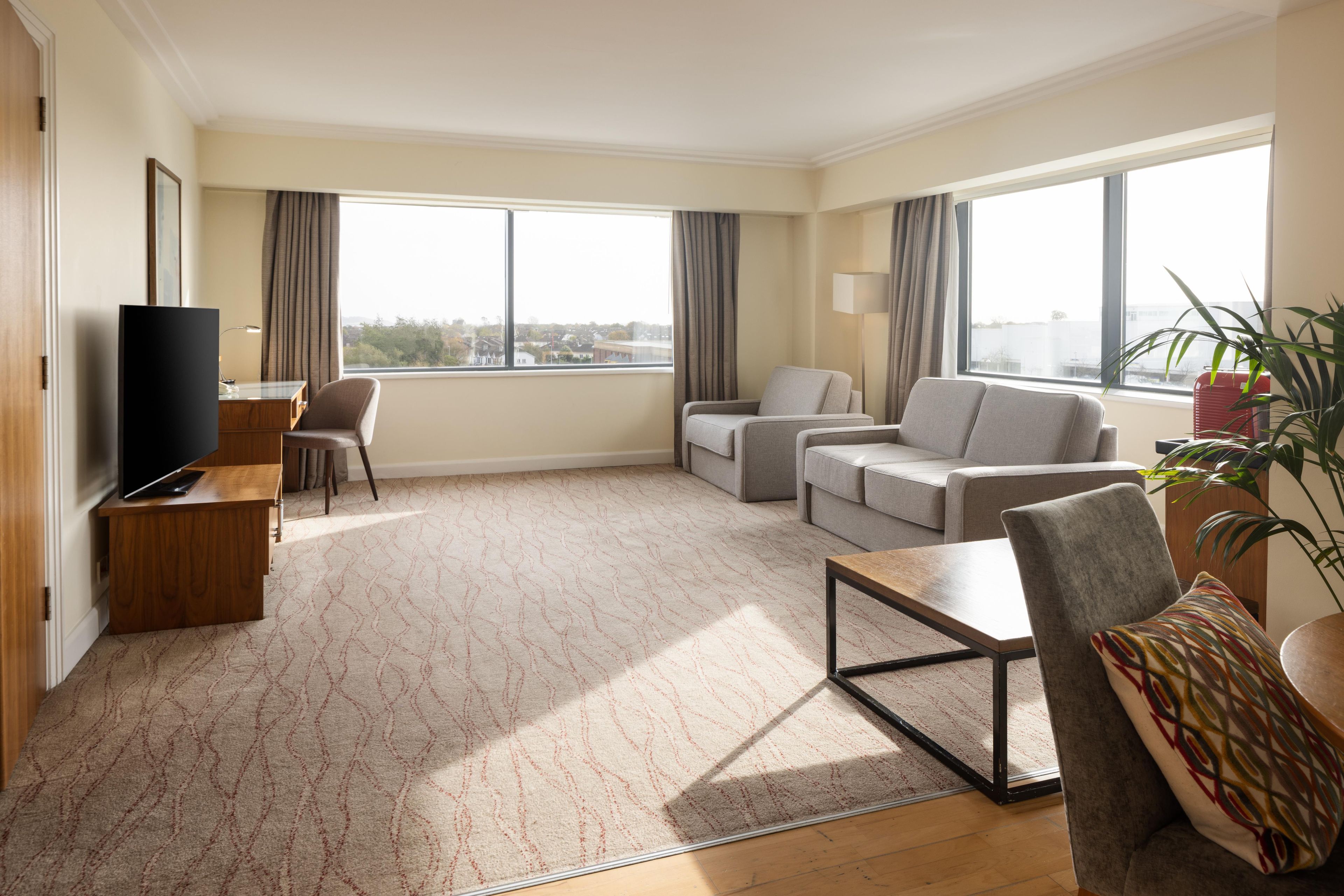 A modern hotel room with a flat-screen TV, a sofa, a chair, a coffee table, and large windows offering a city view.