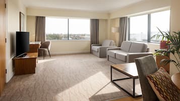 A modern hotel room with a flat-screen TV, a sofa, a chair, a coffee table, and large windows offering a city view.