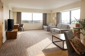 Una habitación de hotel moderna con una televisión de pantalla plana, un sofá, una silla, una mesita de café y ventanas amplias que ofrecen vistas a la ciudad.