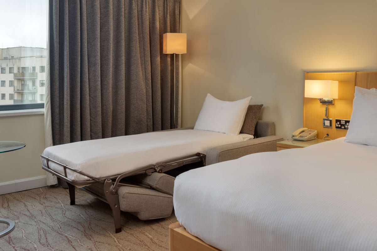 Une chambre d’hôtel avec deux lits, une table de chevet avec une lampe, un téléphone et une fenêtre avec des rideaux.