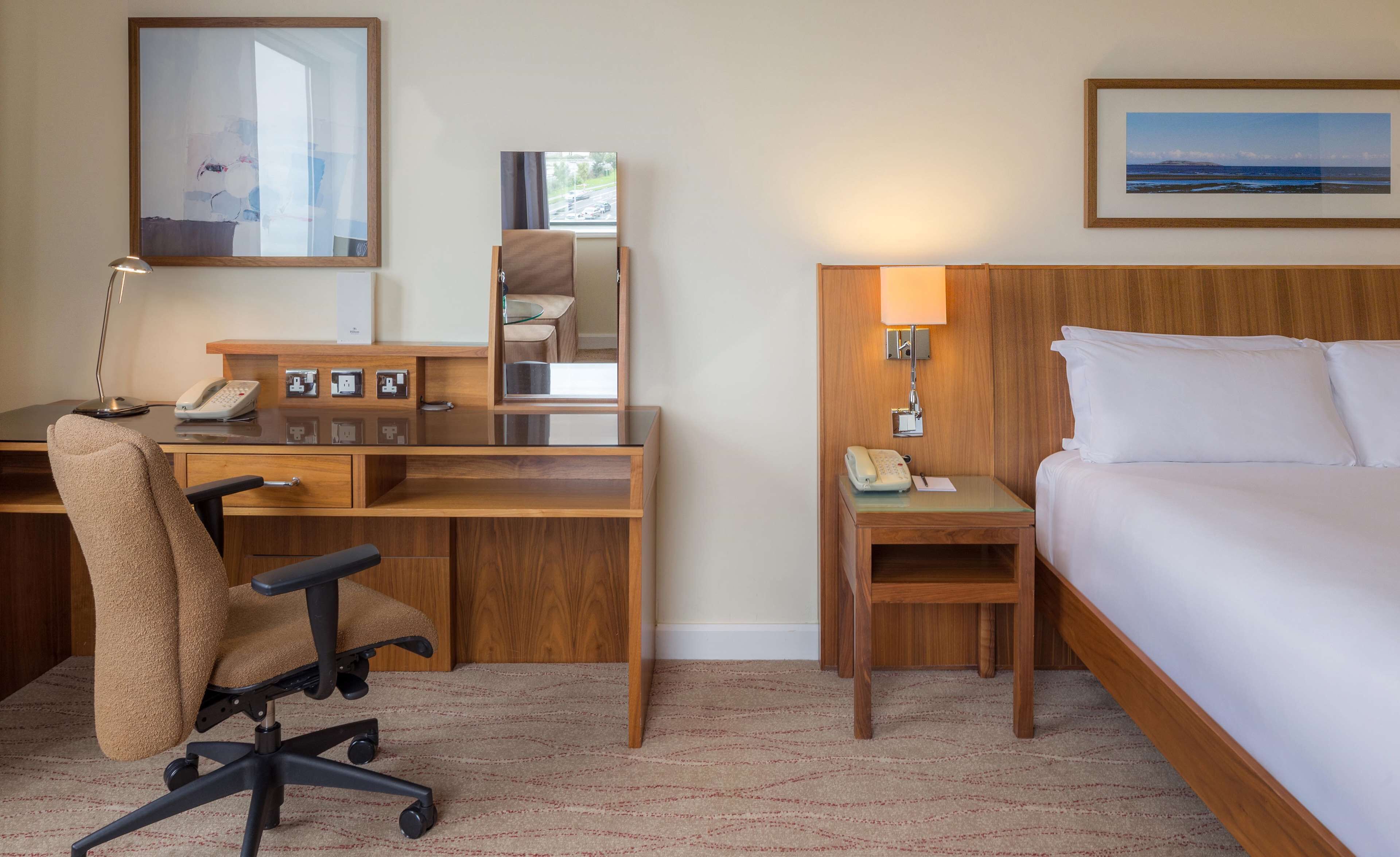 Habitación de hotel con un escritorio de madera, una silla, una cama, una mesita de noche, una lámpara y un cuadro en la pared.