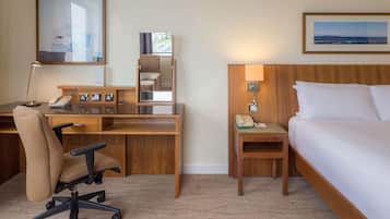 Habitación de hotel con un escritorio de madera, una silla, una cama, una mesita de noche, una lámpara y un cuadro en la pared.