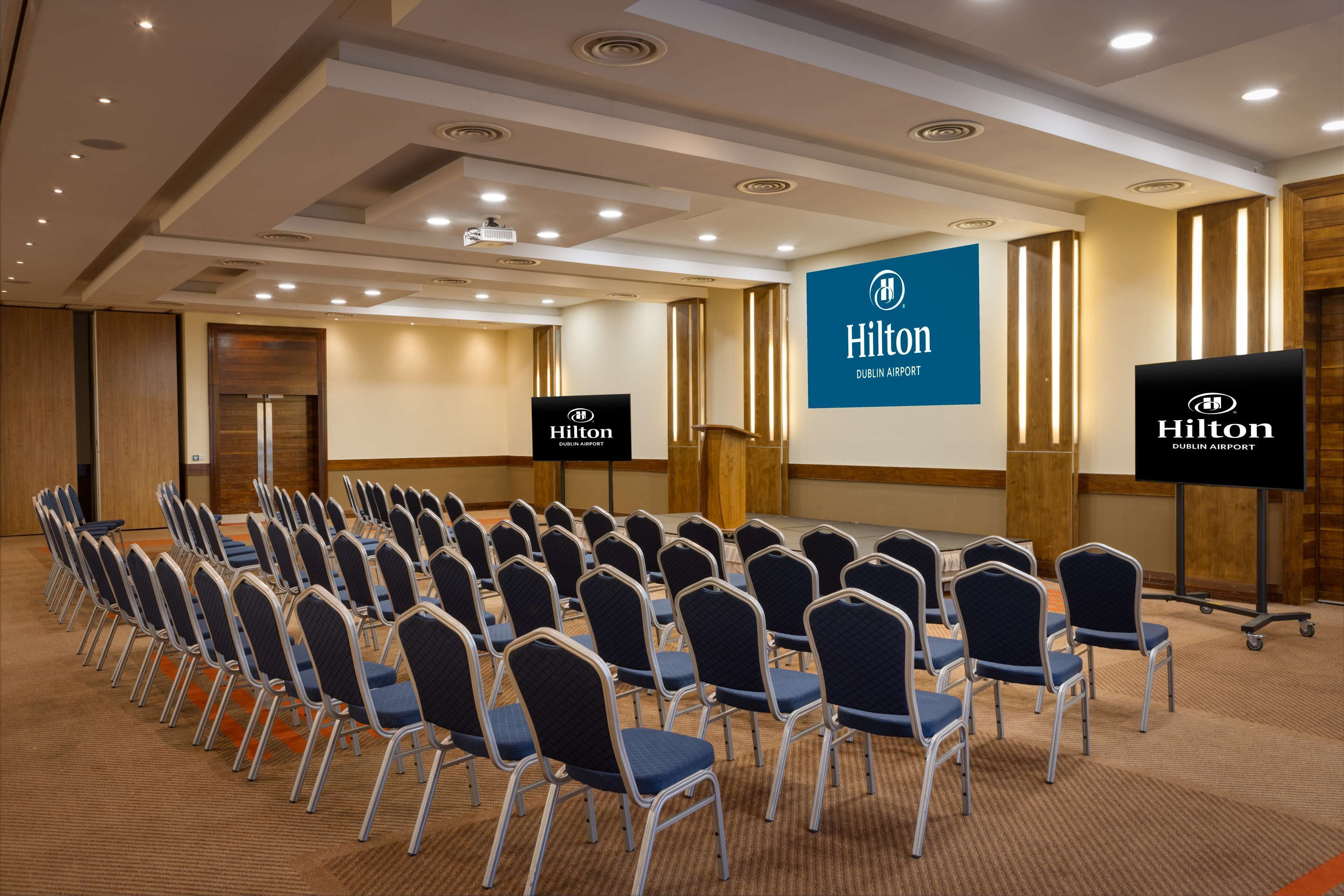 Une salle de conférence avec des rangées de chaises, un pupitre et un écran affichant le logo de l’hôtel Hilton Dublin Airport.