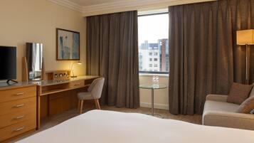 A hotel room with a bed, a desk, a chair, a small table with bottles, a TV, and a window with curtains.