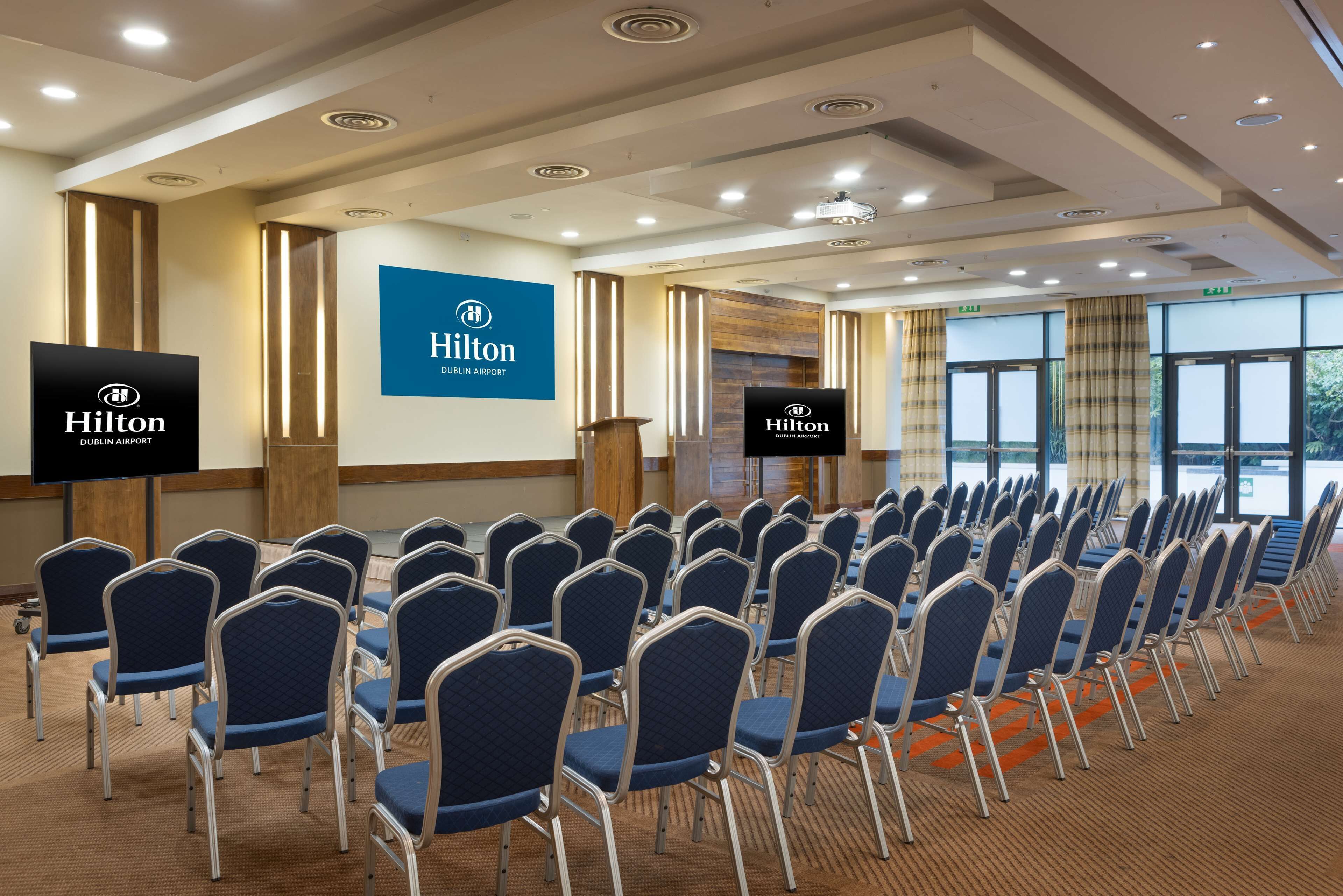A conference room with rows of blue chairs, a podium, and a large screen displaying the Hilton logo.