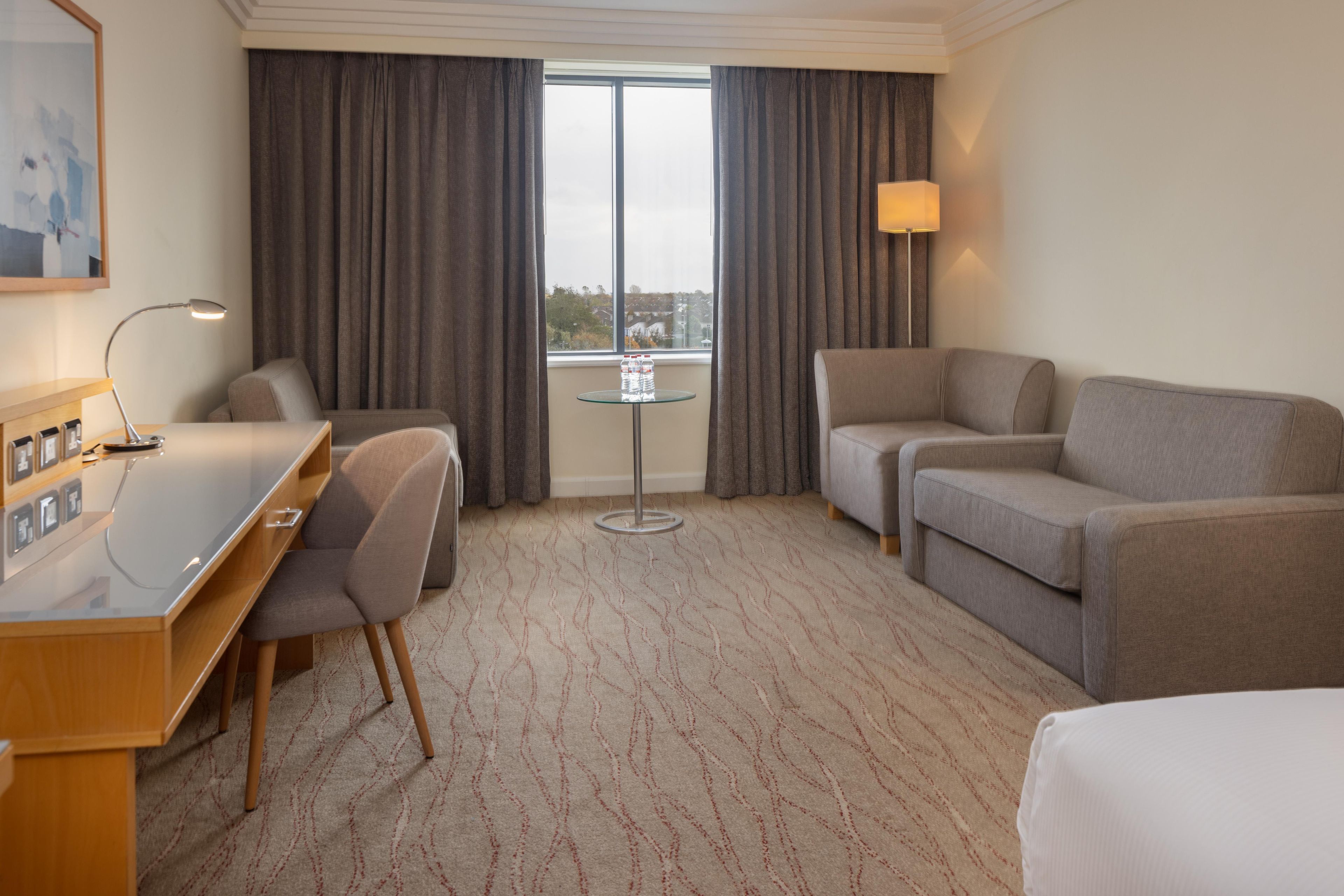 A hotel room with a desk, two chairs, a sofa, a small table with water bottles, and a window with curtains.