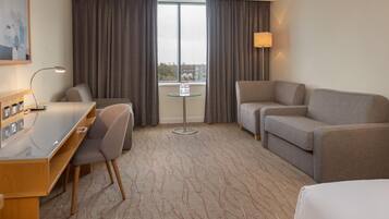 A hotel room with a desk, two chairs, a sofa, a small table with water bottles, and a window with curtains.