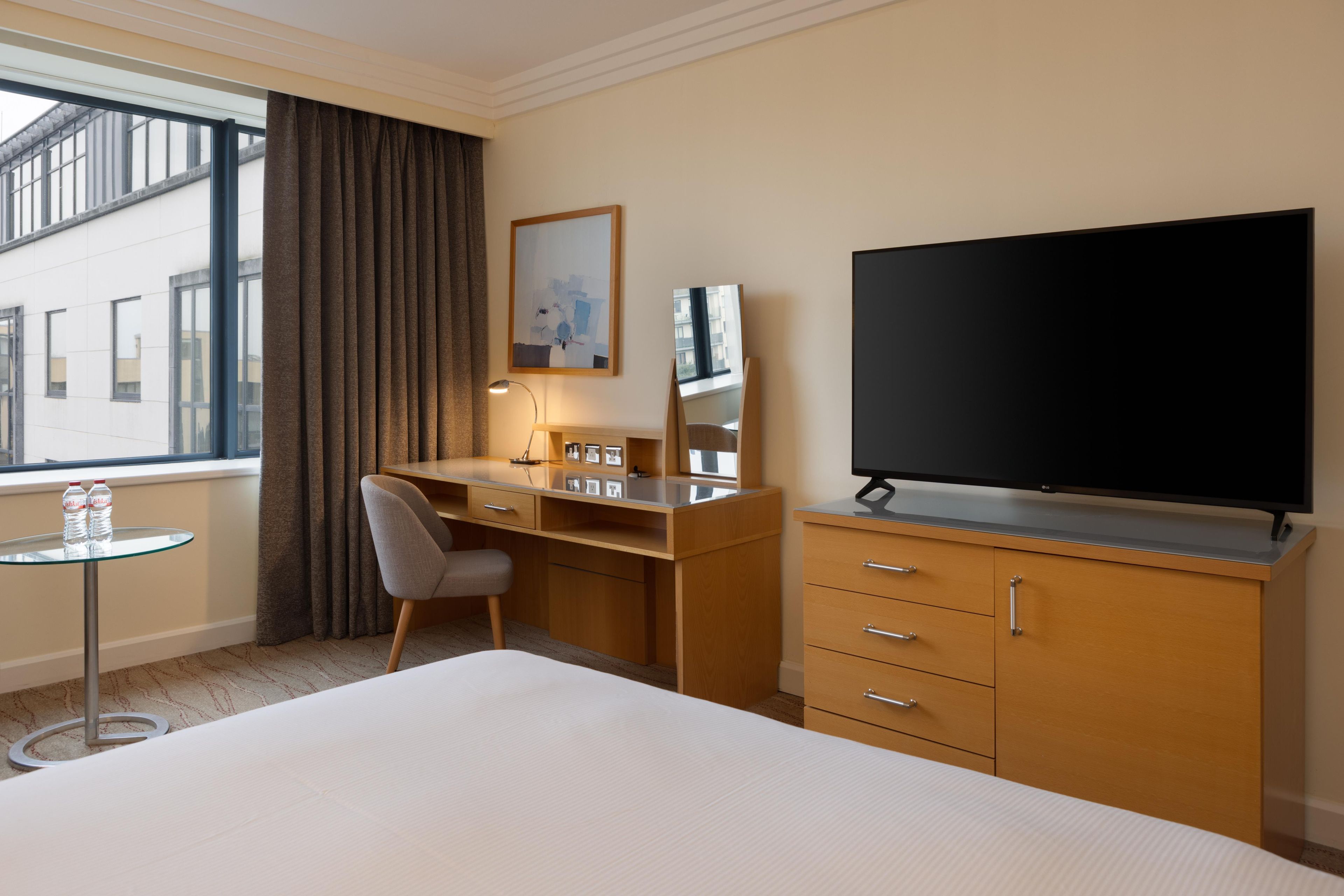 A hotel room with a bed, a desk with a chair, a TV, a small table with two bottles, and a window with curtains.