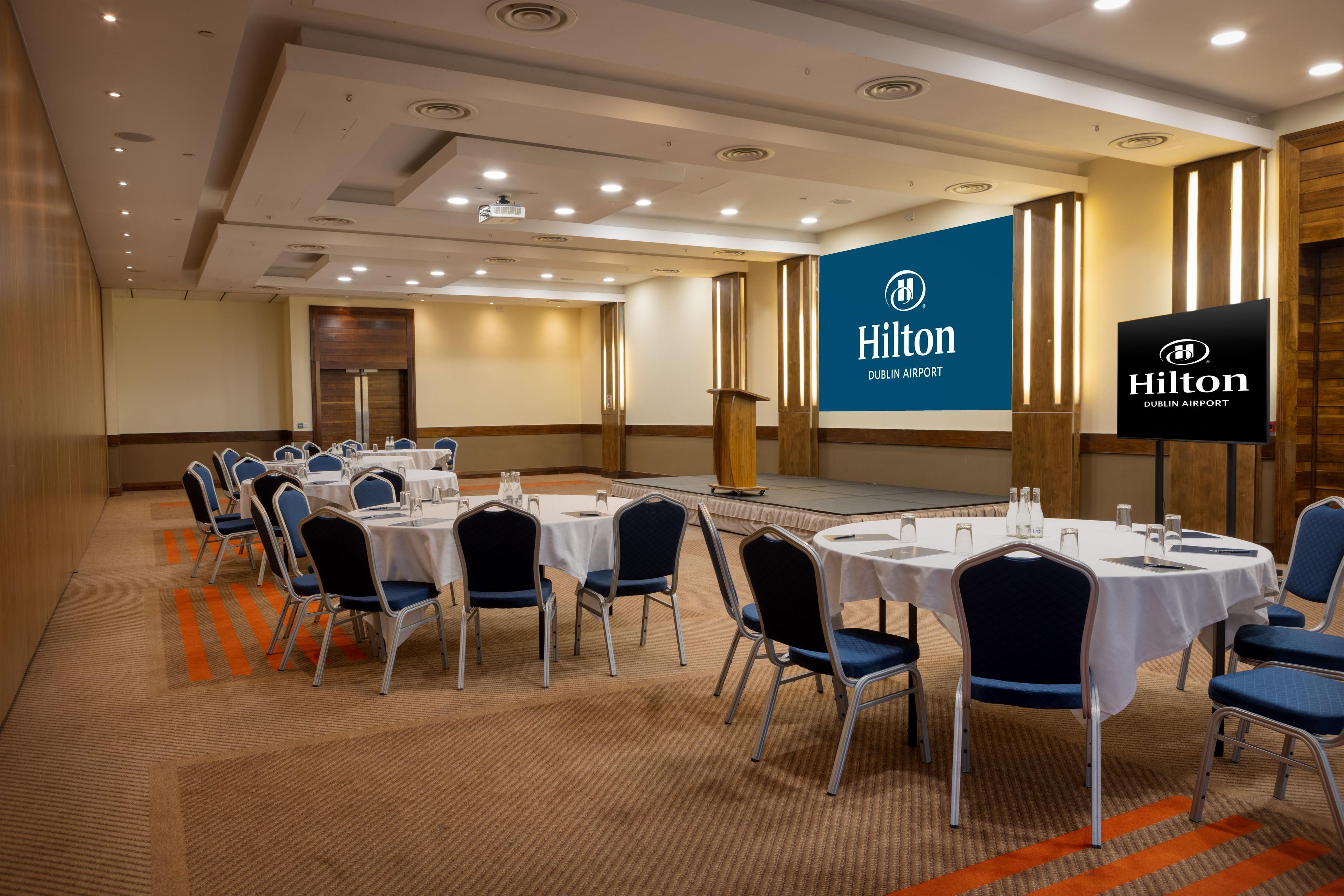 Een congreszaal met ronde tafels, stoelen en een podium, met branding van Hilton Dublin Airport.