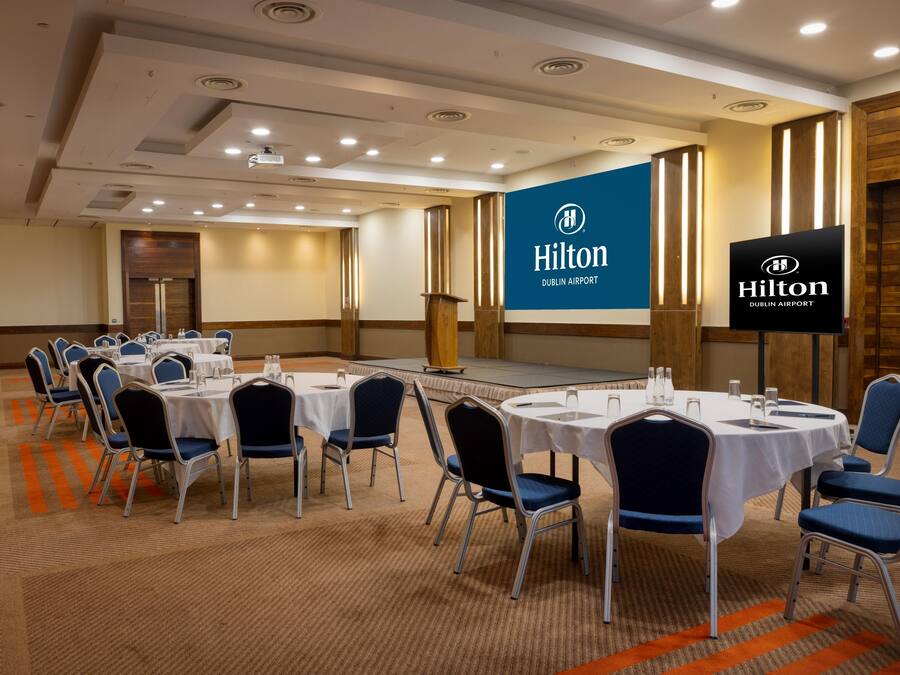 Una sala de conferències amb taules rodones, cadires i un escenari, amb la marca Hilton Dublin Airport.