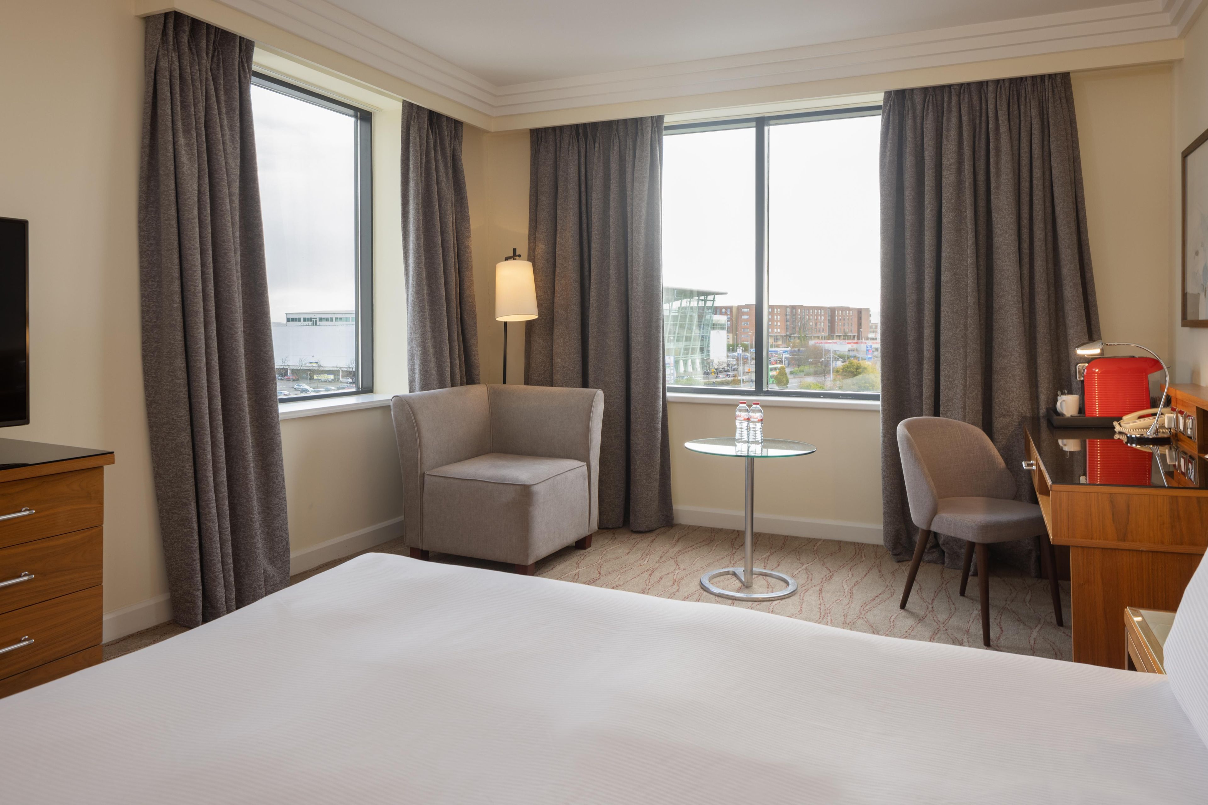 Hotellihuone, jossa on sänky, työpöytä, tuoli, lamppu ja suuret ikkunat, joista on näkymä kaupunkiin.