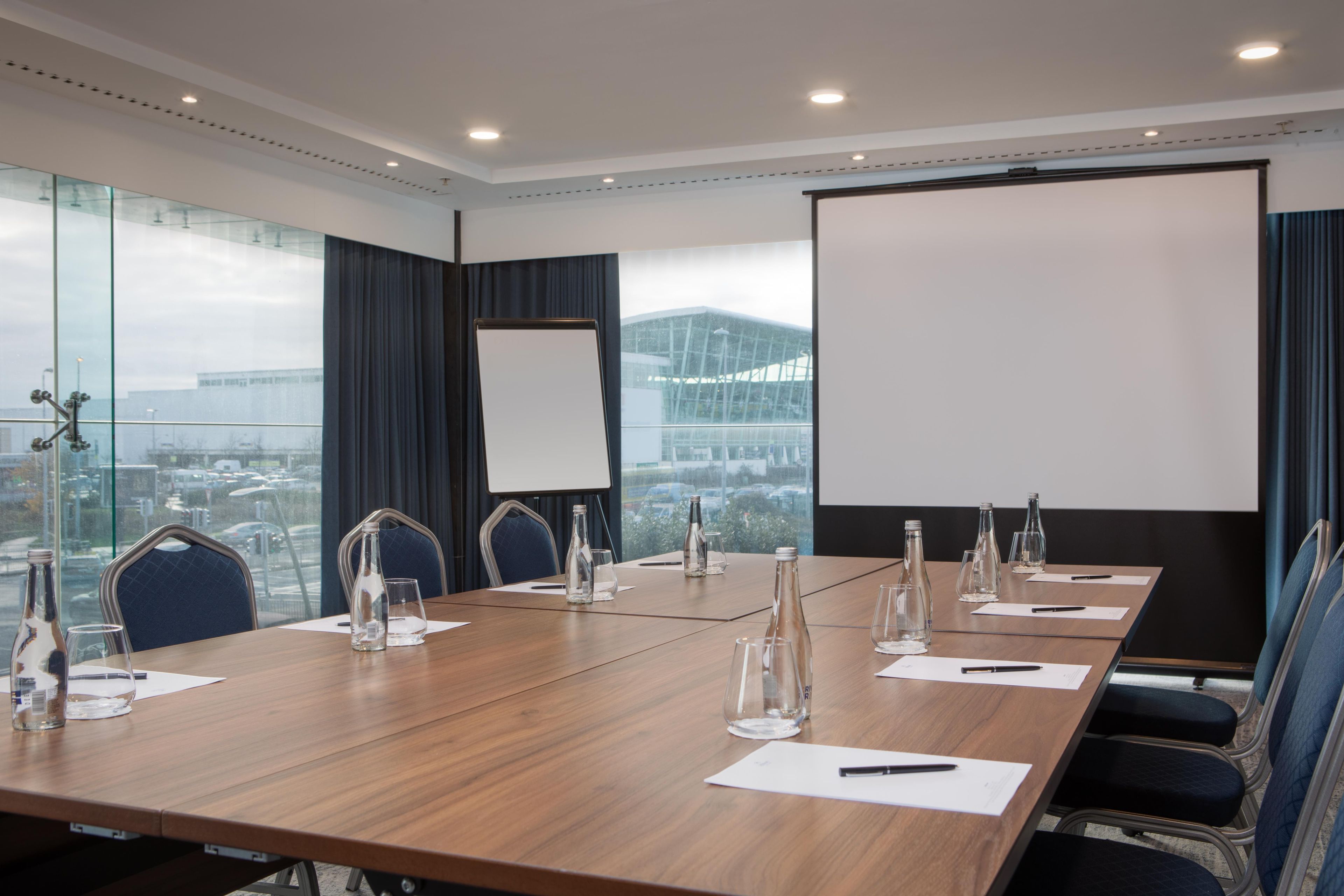 Ein Konferenzraum mit langem Tisch, Stühlen, einer Projektionsleinwand und Blick auf ein Gebäude durch große Fenster.