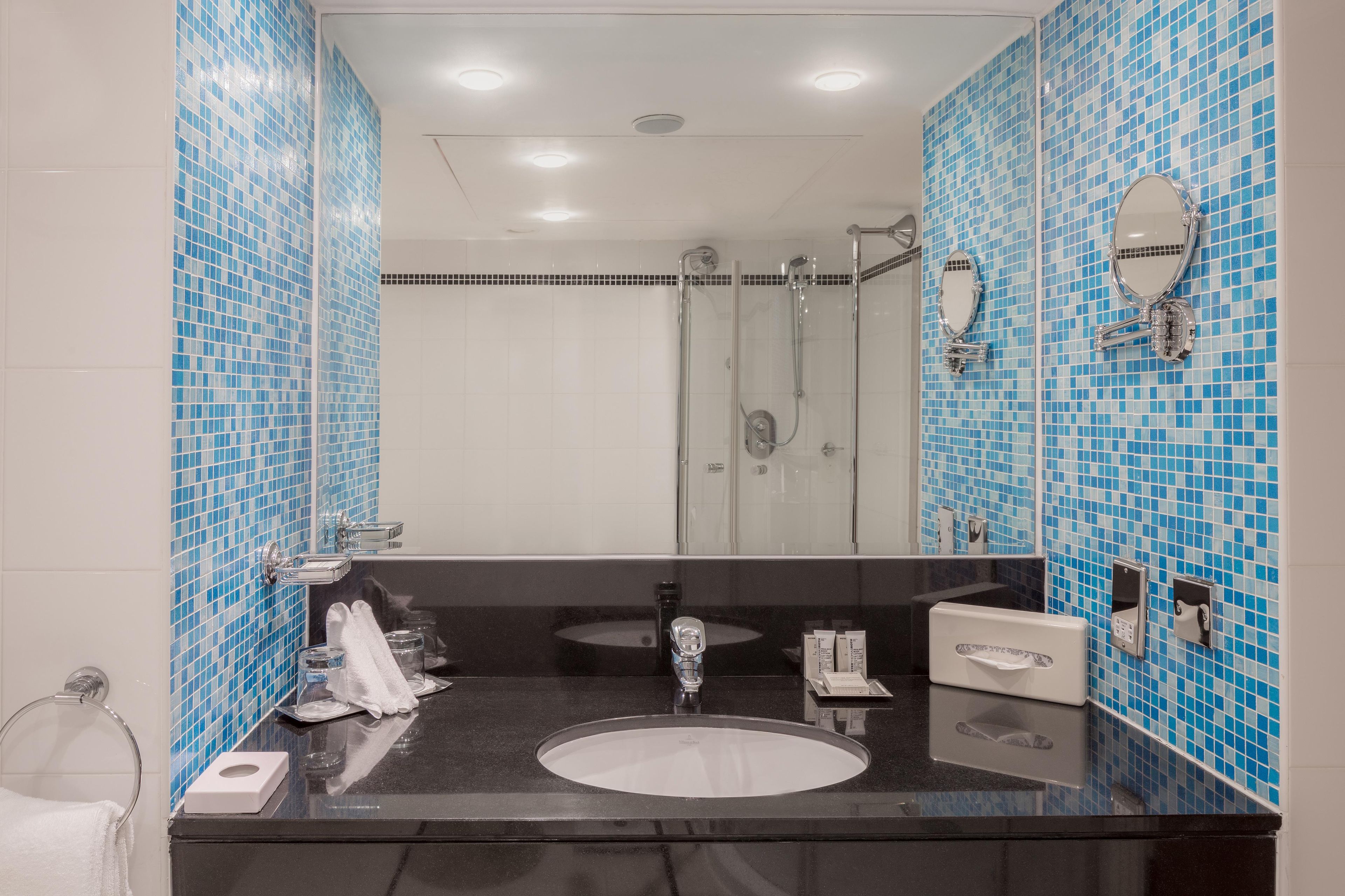 A bathroom with a large mirror, a sink, a shower, and blue-tiled walls.