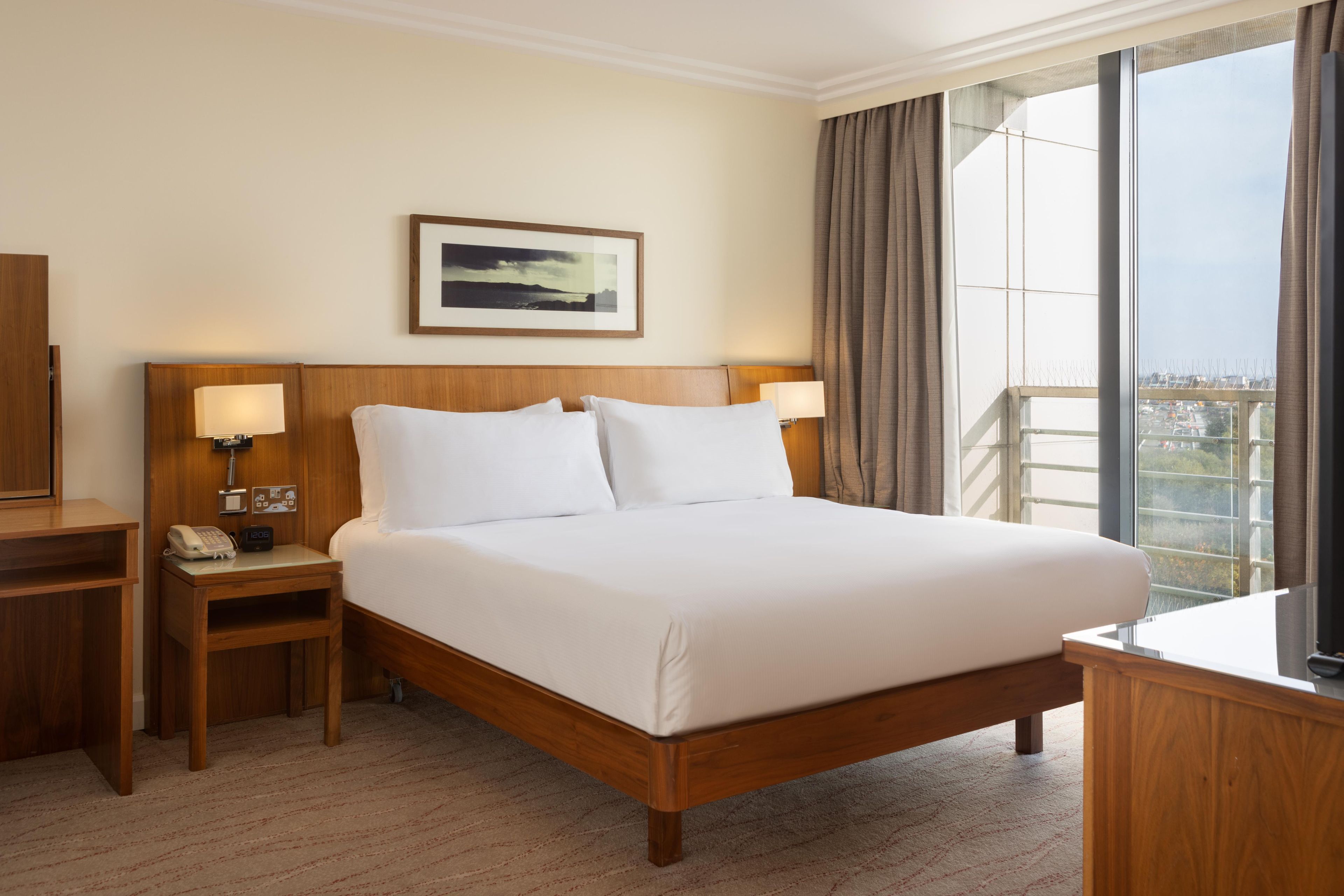 Quarto de hotel com uma cama grande, duas mesas de cabeceira, um criado-mudo, telefone, relógio, quadro na parede e vista para a cidade pela janela.