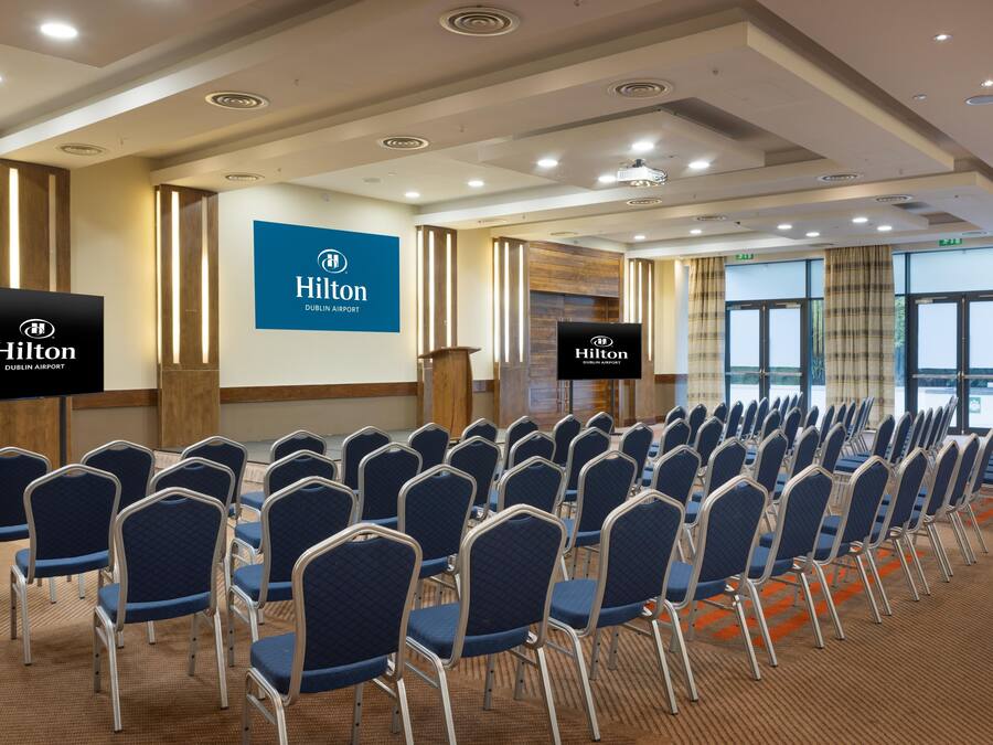 A conference room with rows of chairs, a podium, and a large screen displaying the Hilton Dublin Airport logo.