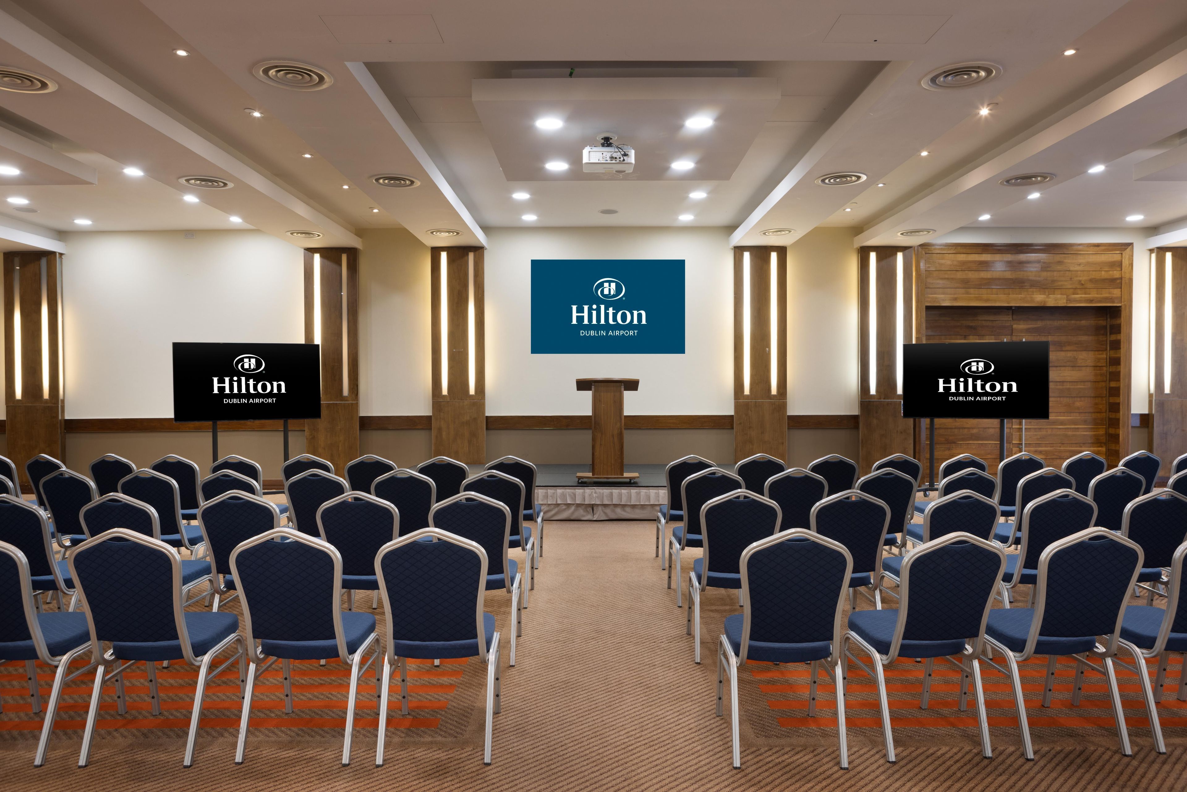 Een vergaderruimte met rijen stoelen, een spreekgestalte en een groot scherm waarop het logo van Hilton Dublin Airport staat.
