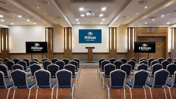 Ein Konferenzraum mit Reihen von Stühlen, einem Rednerpult und einem großen Bildschirm, der das Logo des Hilton Dublin Airport zeigt.