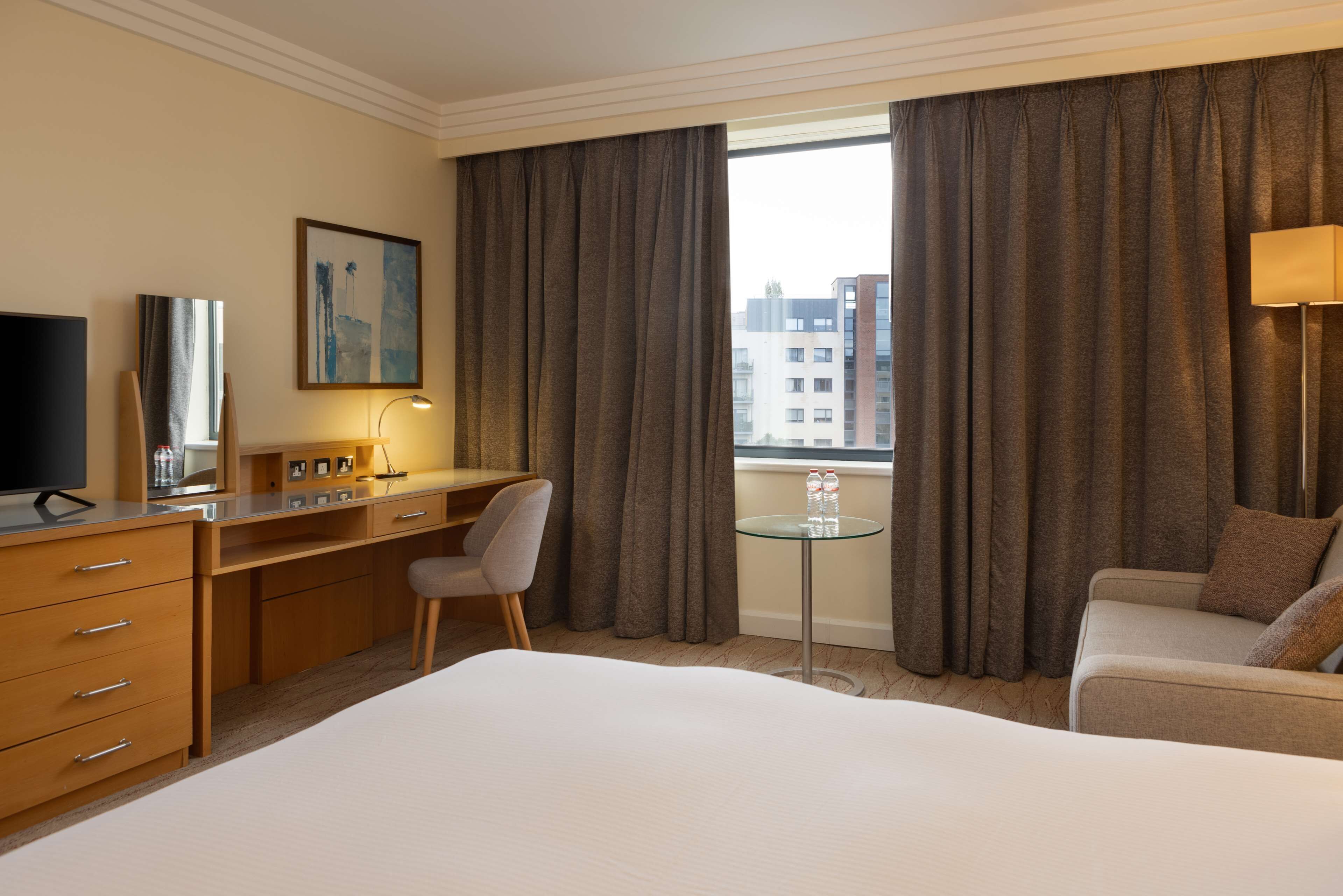 A hotel room with a bed, a desk, a chair, a small table with a lamp, and a window with curtains.