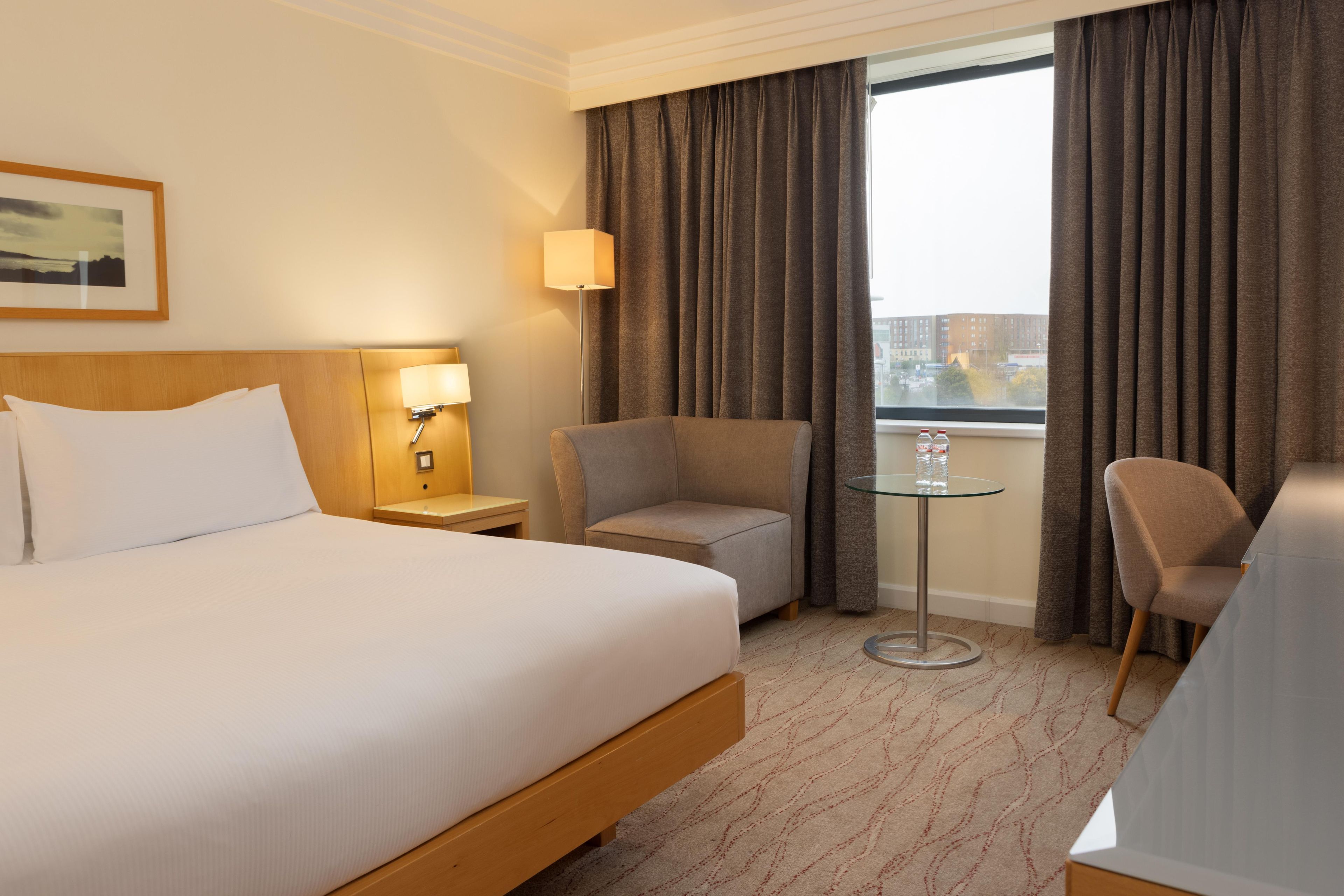 A hotel room with a bed, a desk, two chairs, a small table, and a window with curtains.