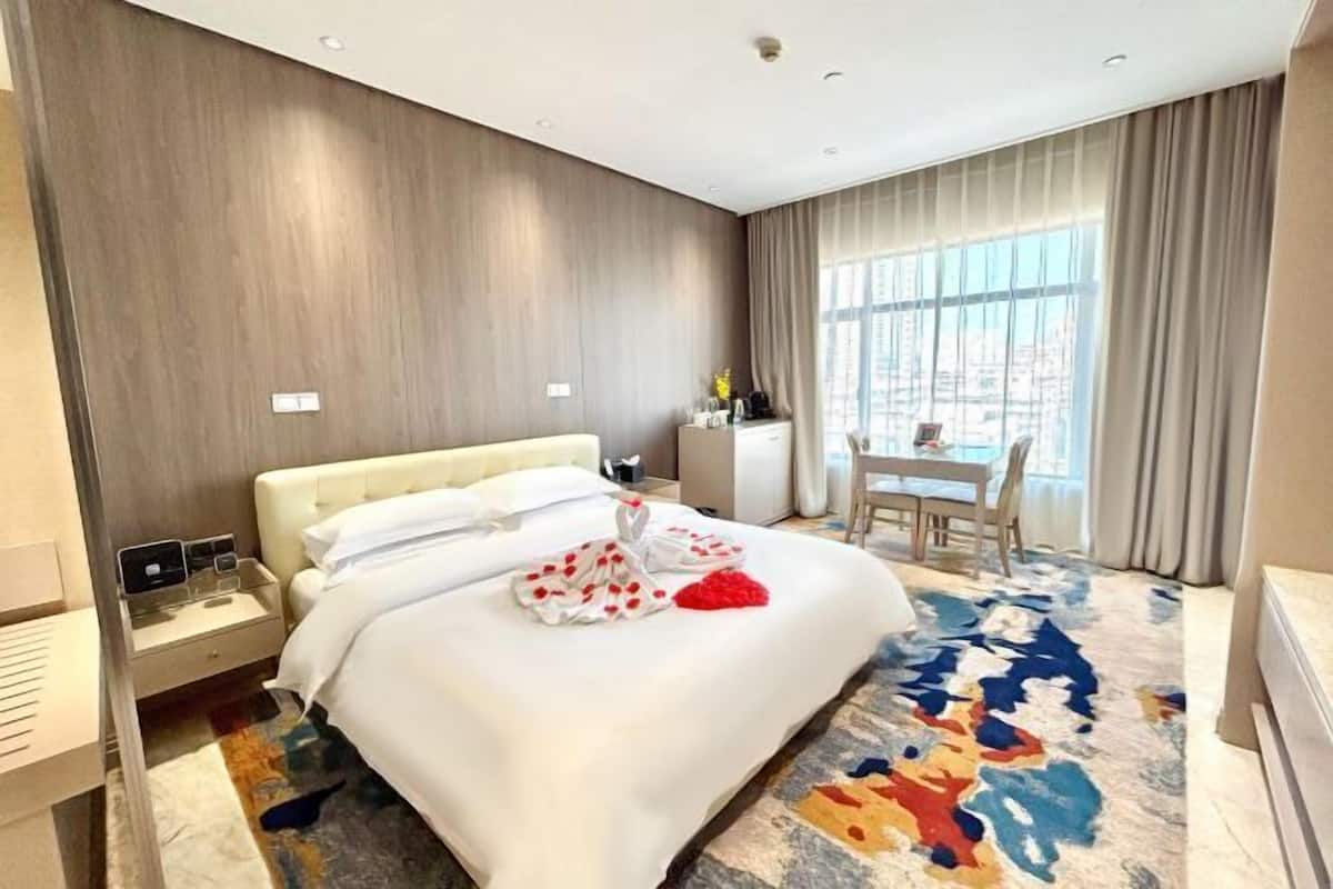 Habitación de hotel con cama, mesita de noche y vistas de la ciudad a través de amplios ventanales.