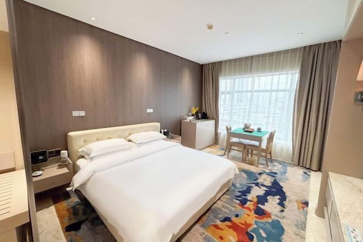 Habitación de hotel con una cama grande, un escritorio y una silla. Se ve una ventana con cortinas y vista a la ciudad.