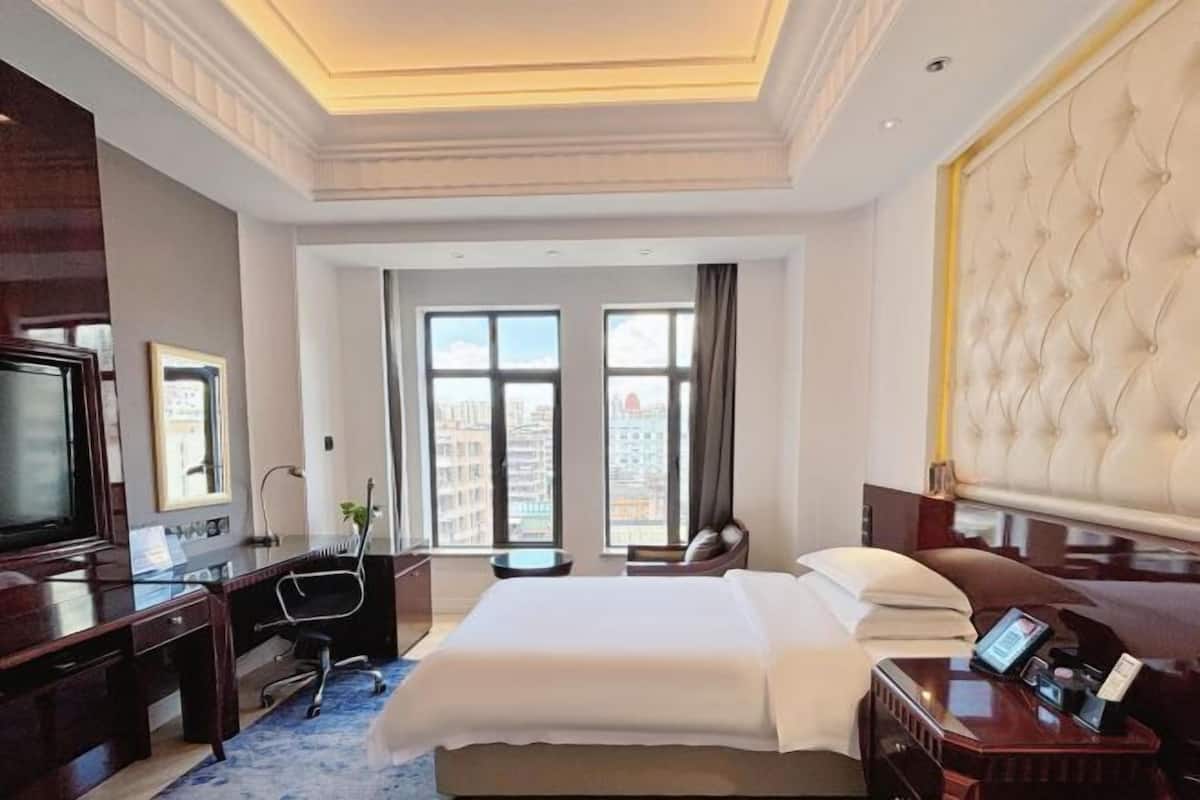 Habitación de hotel con una cama grande, un escritorio con una silla, televisión y vistas de la ciudad a través de amplias ventanas.