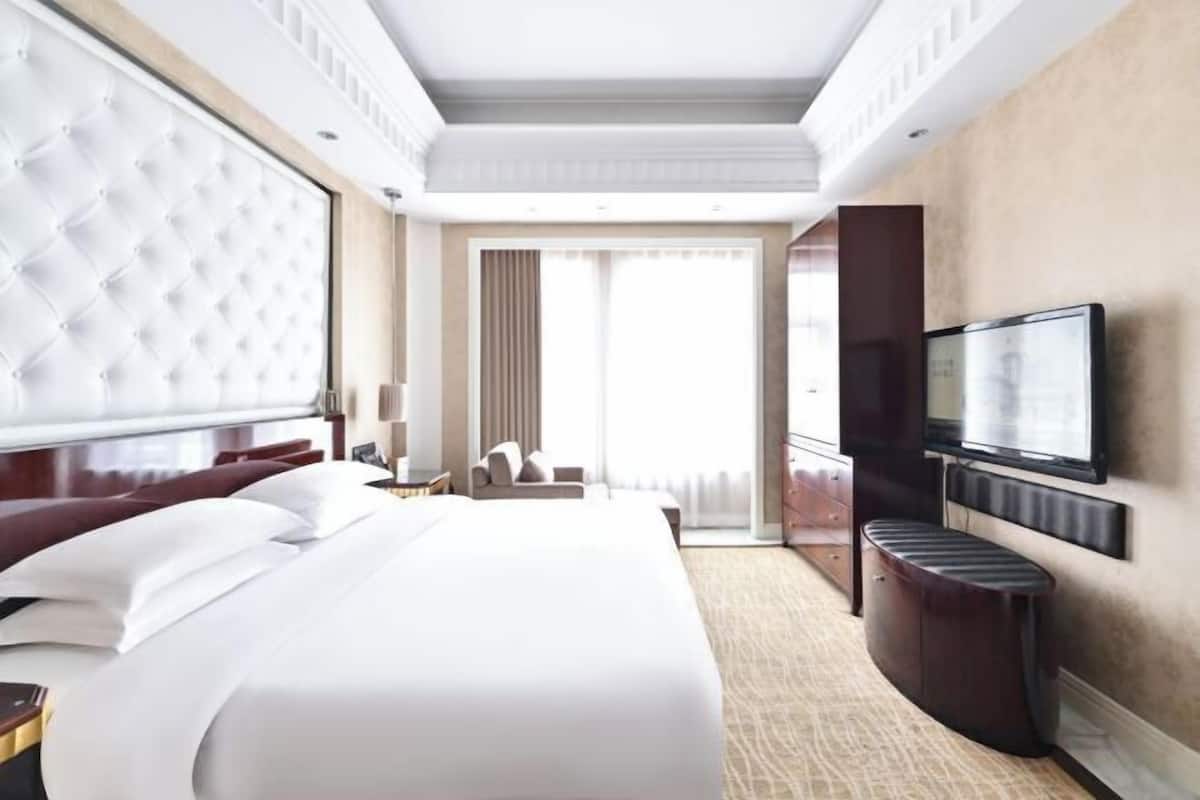 Habitación de hotel con una cama grande, televisión montada en la pared, zona de estar con una silla y una mesita, y un ventanal con cortinas.