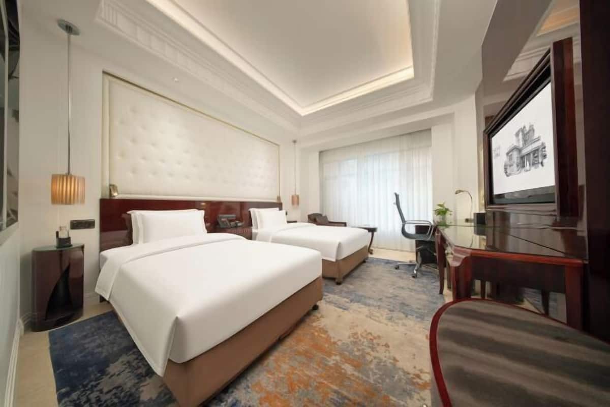 Habitación de hotel con dos camas, un escritorio, una silla, un televisor y un ventanal con cortinas.