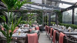 Un espacio exterior para comer con mesas y sillas, rodeado de follaje y un recinto de cristal.