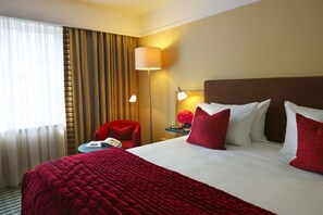 A hotel room with a large bed, red bedding, a red armchair, a bedside table with a lamp, and a view of the outside through curtains.