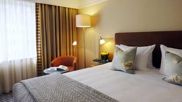 Habitación de hotel con una cama grande, un escritorio con silla, una lámpara y vistas al exterior a través de cortinas.