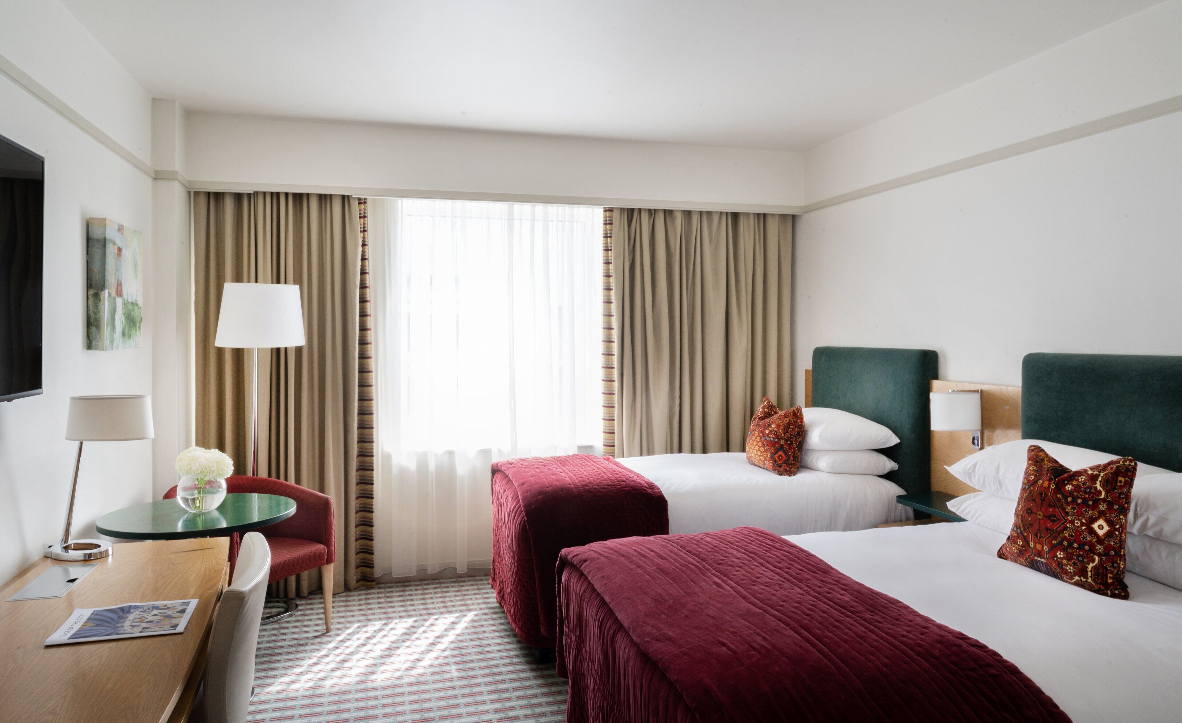 Ein Hotelzimmer mit zwei Betten, einem Schreibtisch mit Stuhl, einer Lampe und einem Fenster mit Vorhängen.
