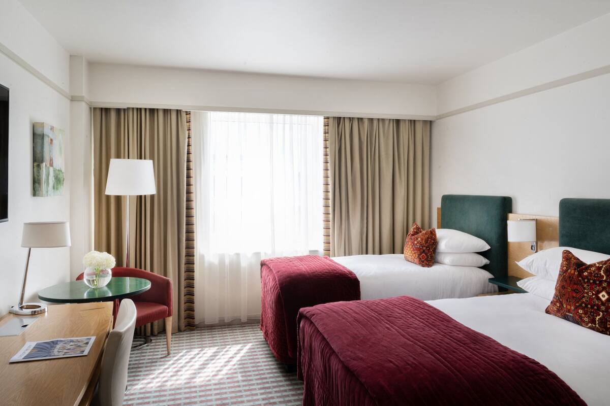 Ein Hotelzimmer mit zwei Betten, einem Schreibtisch mit Stuhl, einer Lampe und einem Fenster mit Vorhängen.