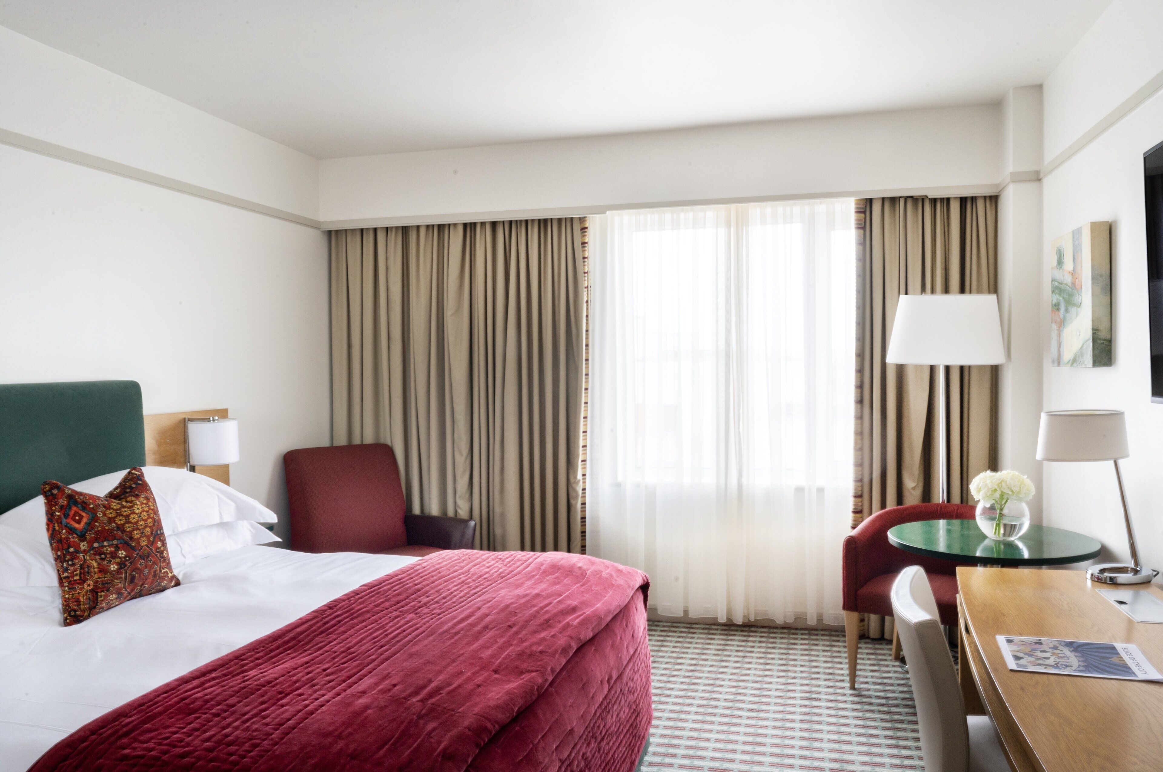 A hotel room with a bed, a desk, a chair, a lamp, and a window with curtains.
