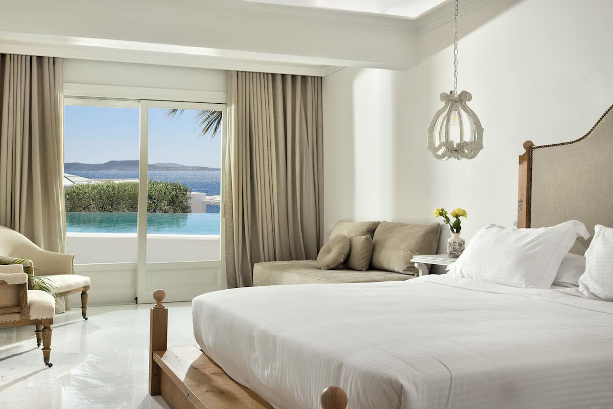 Une chambre avec un grand lit, une chaise, une petite table avec un vase et une vue sur la mer.