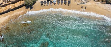 Vista aérea de una playa con aguas turquesas cristalinas, una costa rocosa y una fila de tumbonas.