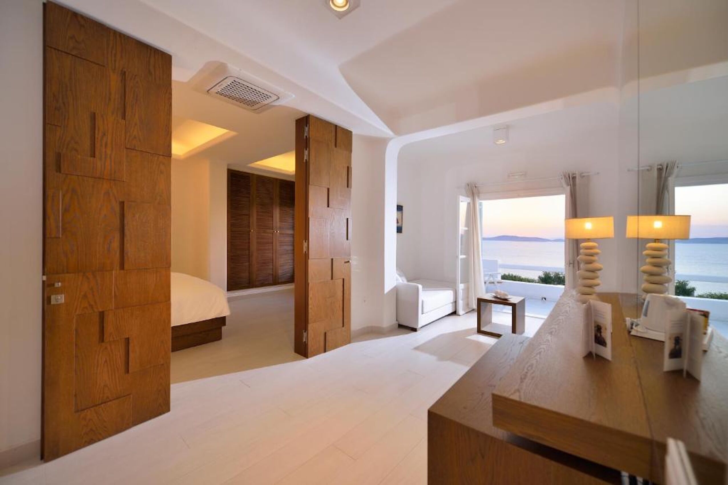 A modern hotel room with a large bed, a wooden wardrobe, a seating area, and a view of the sea.