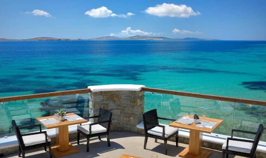 A terrace with tables and chairs overlooking a clear blue sea and distant land.