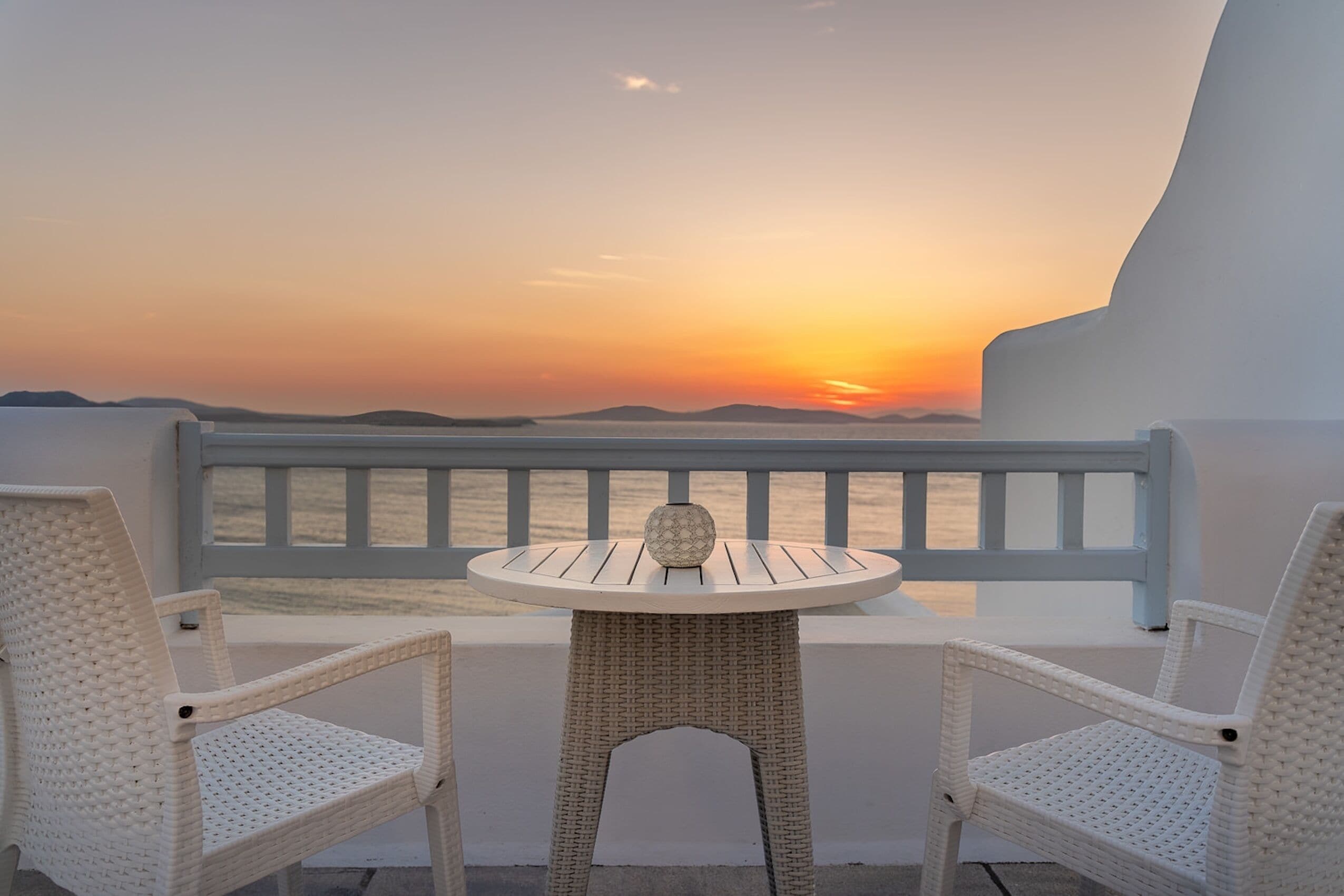 A balcony with a wicker table and two chairs, overlooking a sunset over water.