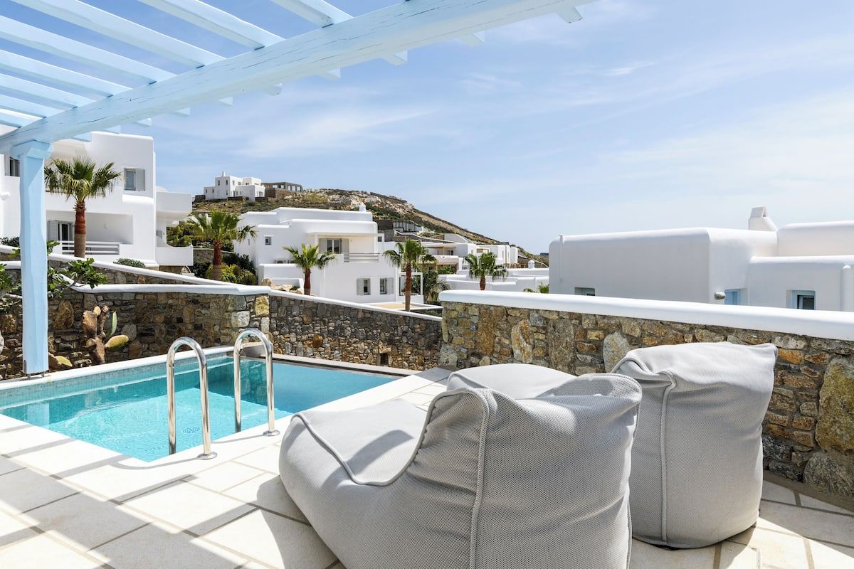 Un espace aménagé au bord de la piscine offrant une vue dégagée sur des bâtiments blancs et un mur de pierre.