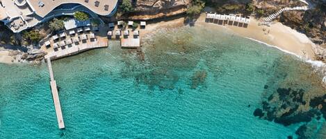 An aerial view of a coastal resort with a clear turquoise sea, a sandy beach, and a pool area with sun loungers.