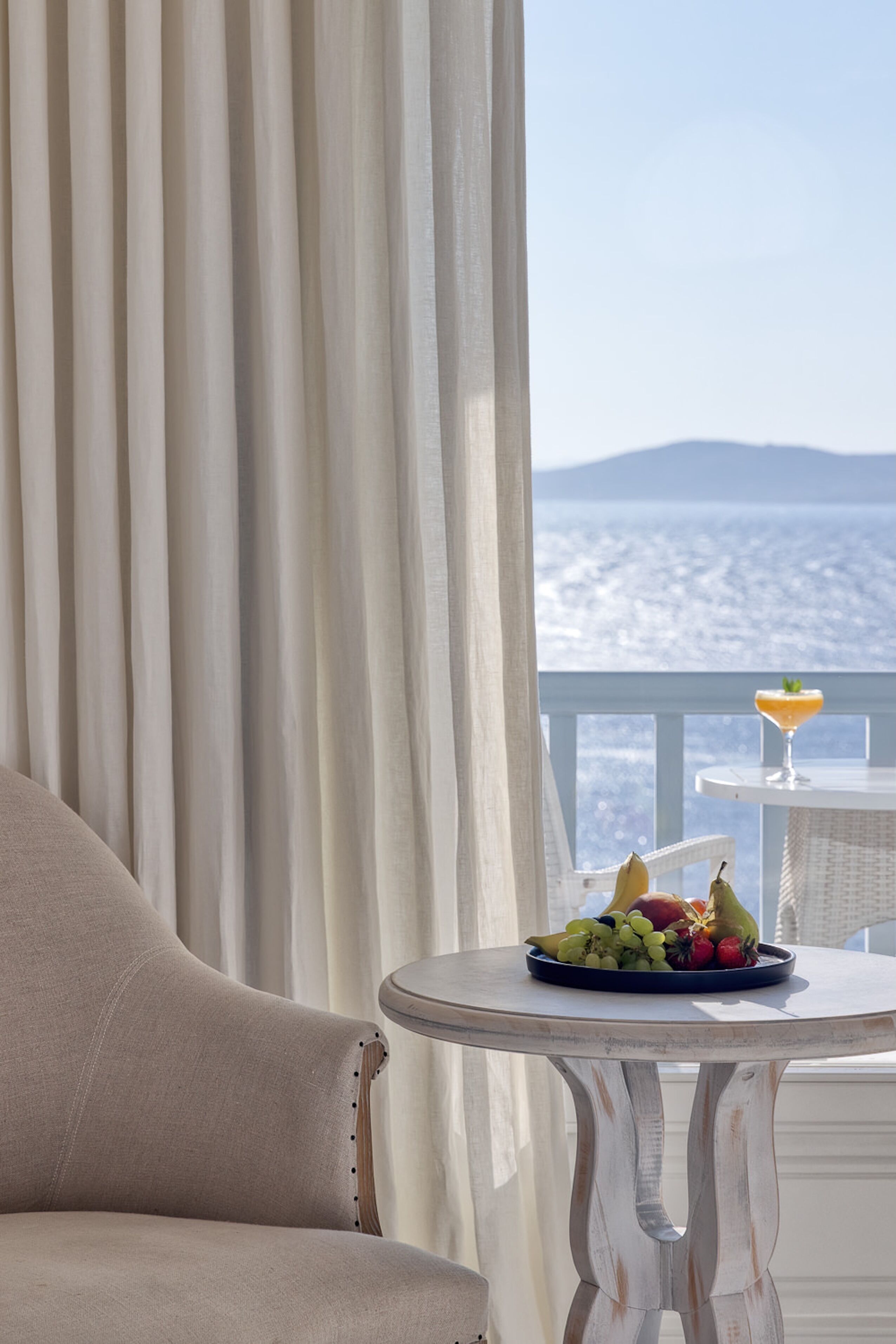 Un balcón con mesa dispuesta, frutas, una bebida, cortinas y vista al mar.
