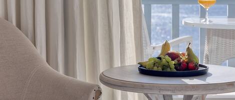 Un balcón con mesa dispuesta, frutas, una bebida, cortinas y vista al mar.