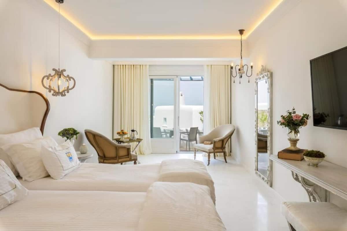 Une chambre d’hôtel avec deux lits, un coin salon avec des fauteuils, une table à manger, un miroir et un balcon avec des rideaux.