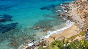 A coastal landscape with clear turquoise waters, rocky shoreline, and a sandy beach.